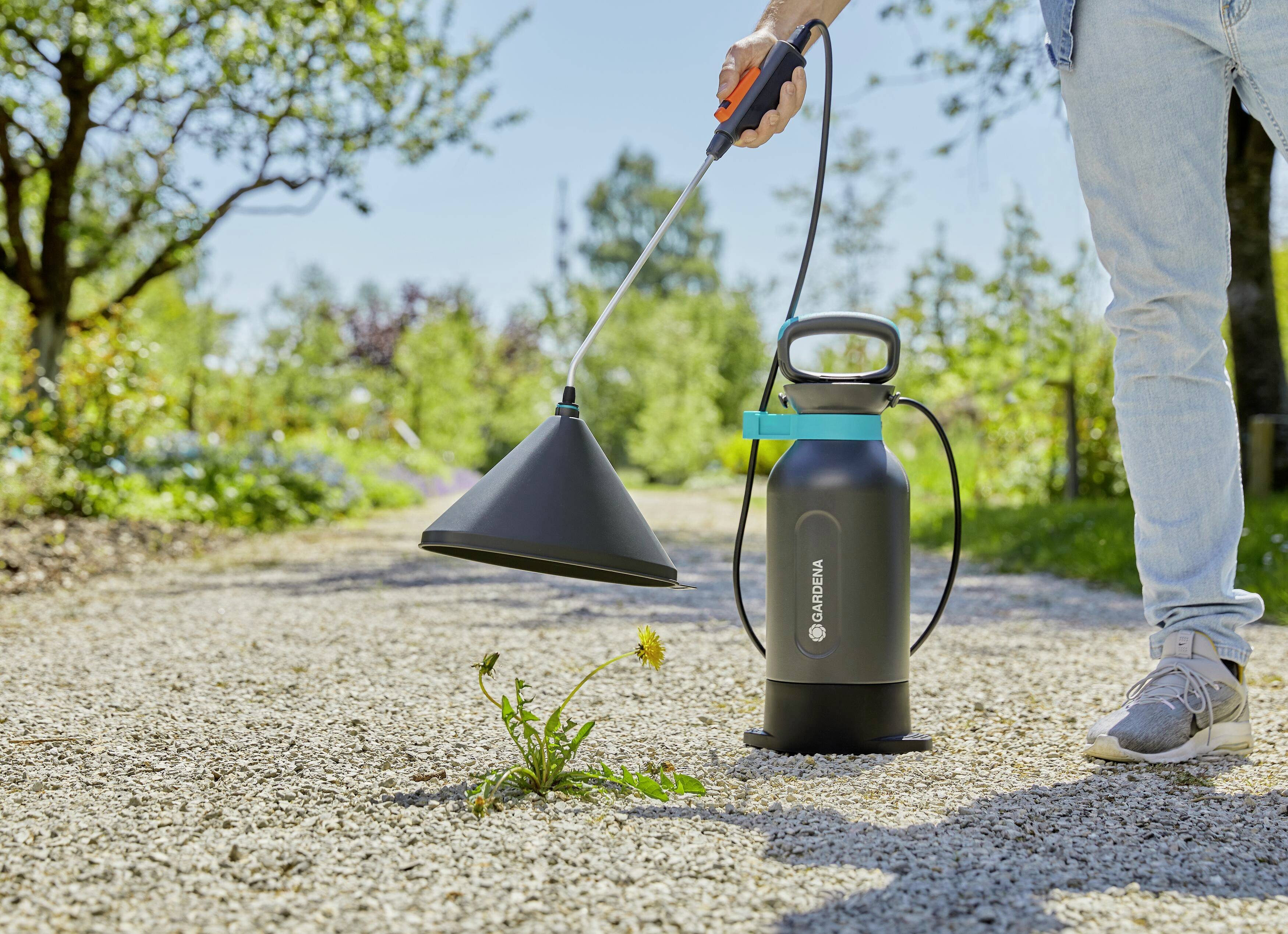 A person is using a gardening tool to remove weeds from a gravel path. Trees and plants are visible in the background.