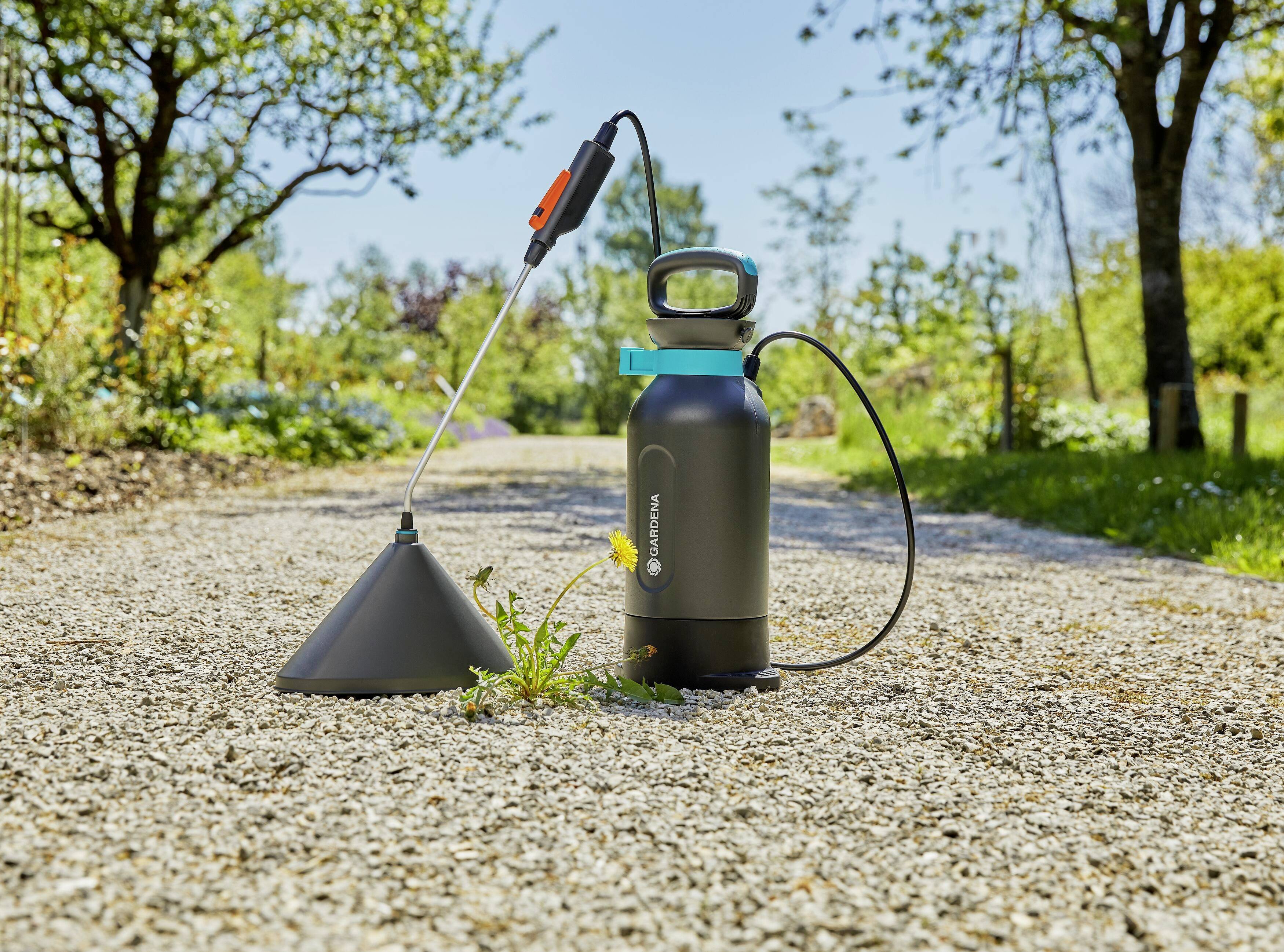 A garden sprayer stands on a gravel path beside a small flowering plant. Trees and shrubs are in the background.