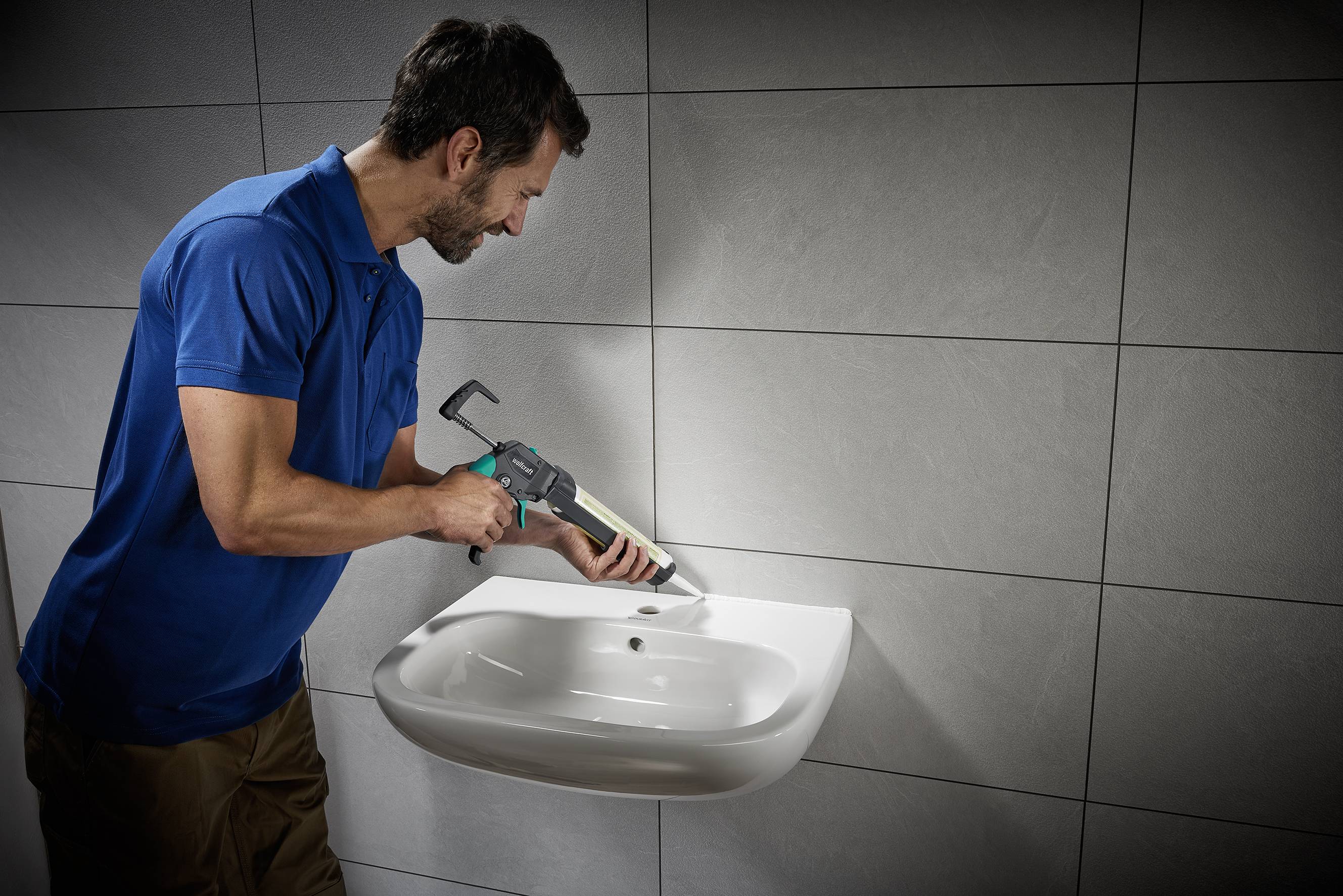 A man in a blue shirt is sealing the edges of a wall-mounted sink in a bathroom using a sealant gun.