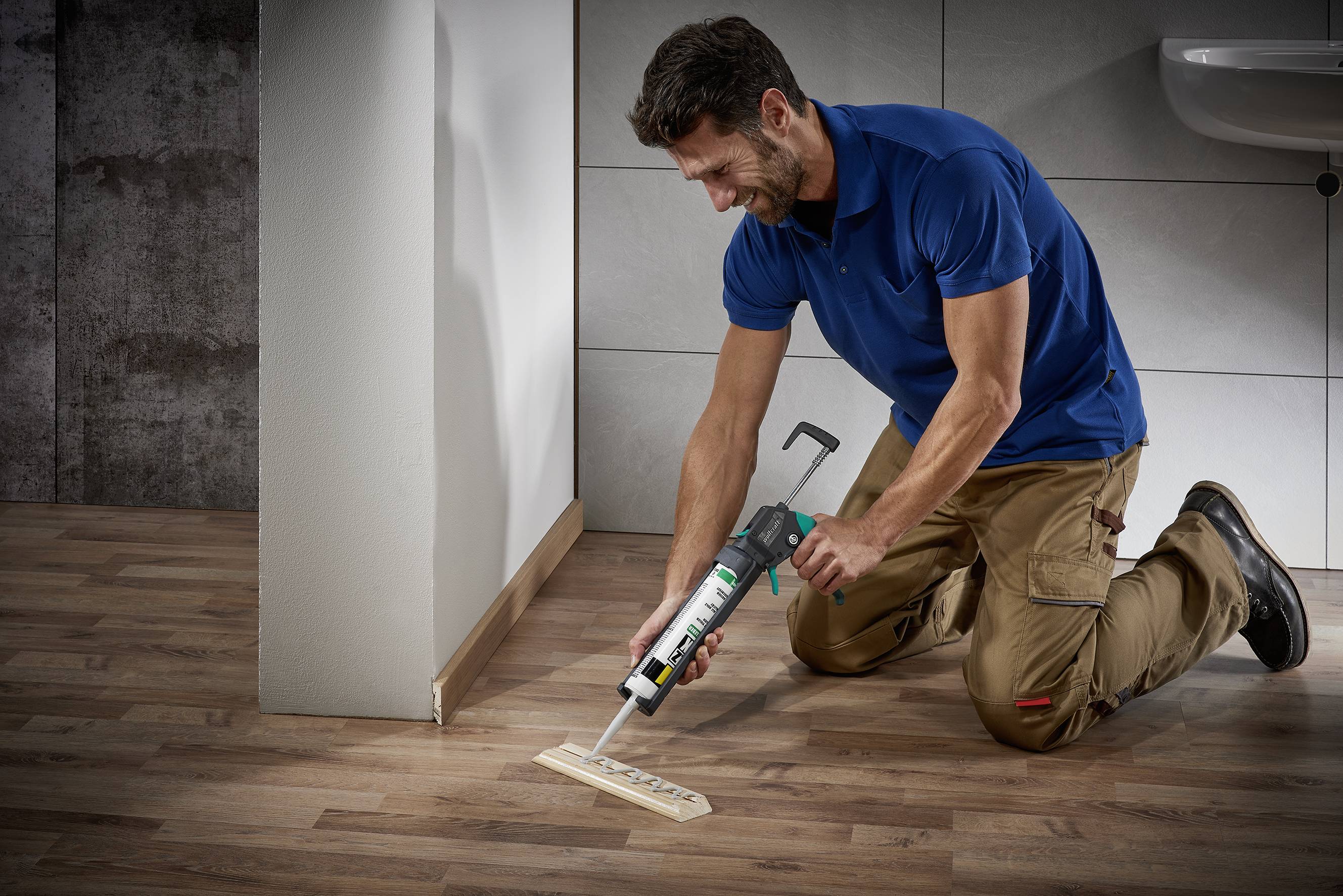 A man in work attire is using a glue gun to secure a piece of wood on a wooden floor.