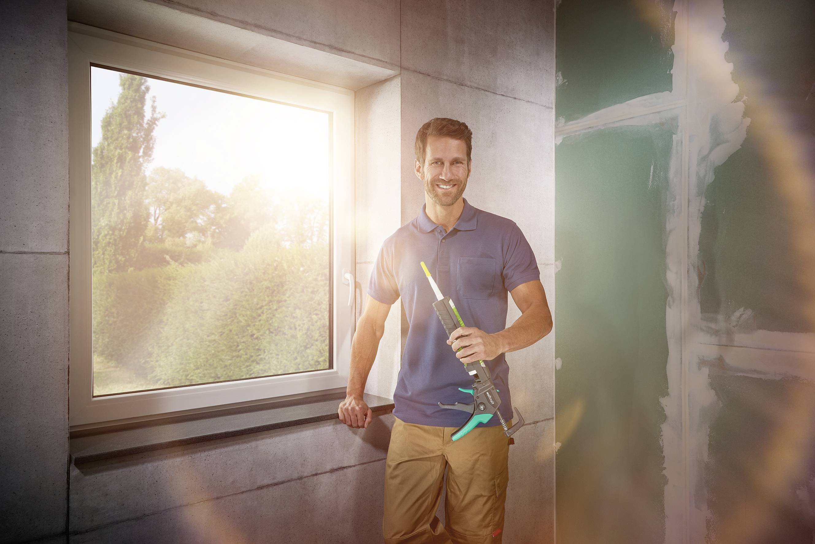 A man in casual clothing stands smiling in a bright room next to a window, holding a caulking gun in his hand.