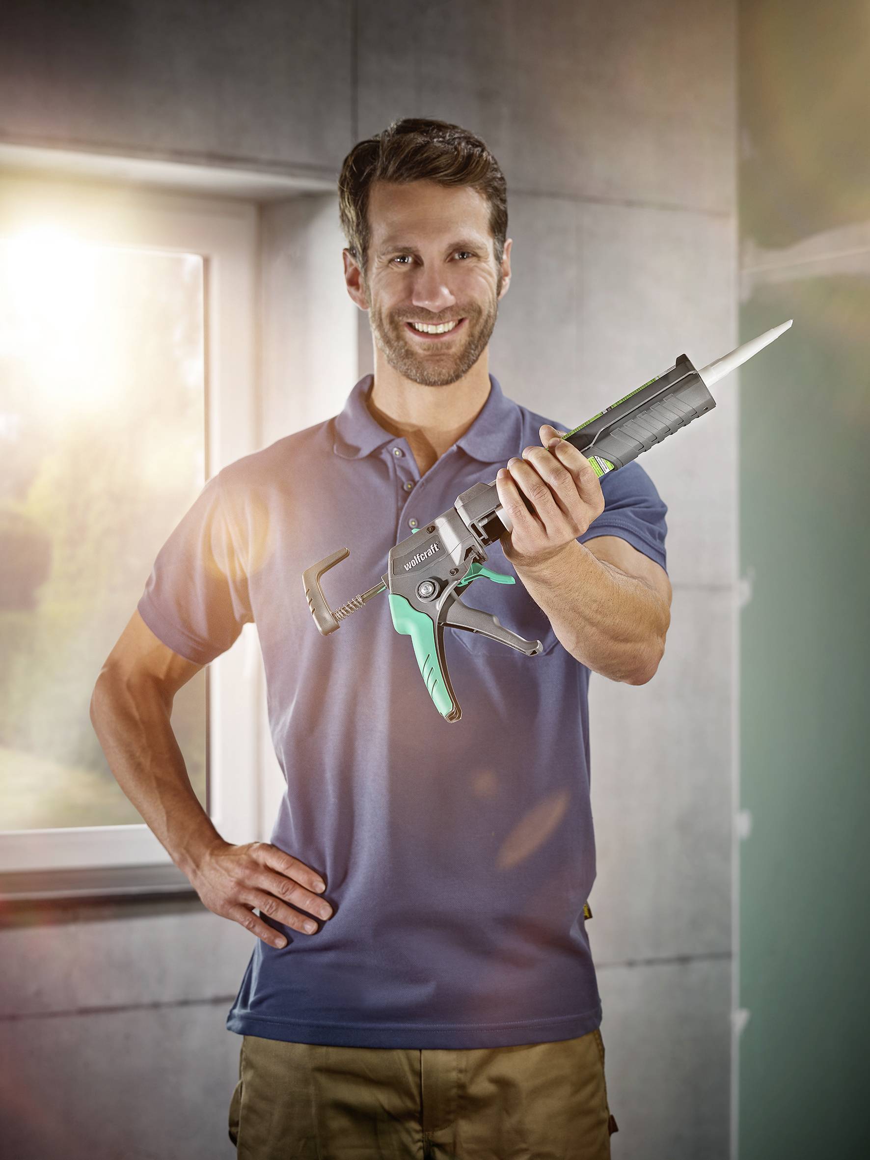 A man in a blue polo shirt is holding a sealant gun and smiling at the camera in a sunlit environment.