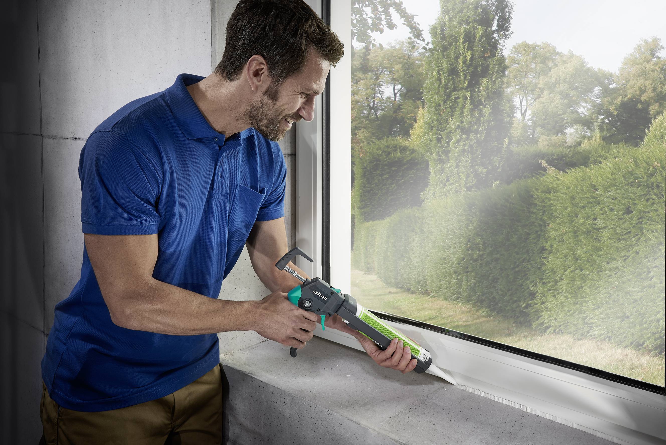 A man is wearing a blue polo shirt and sealing a window with a caulking gun in a sunny room overlooking garden hedges.