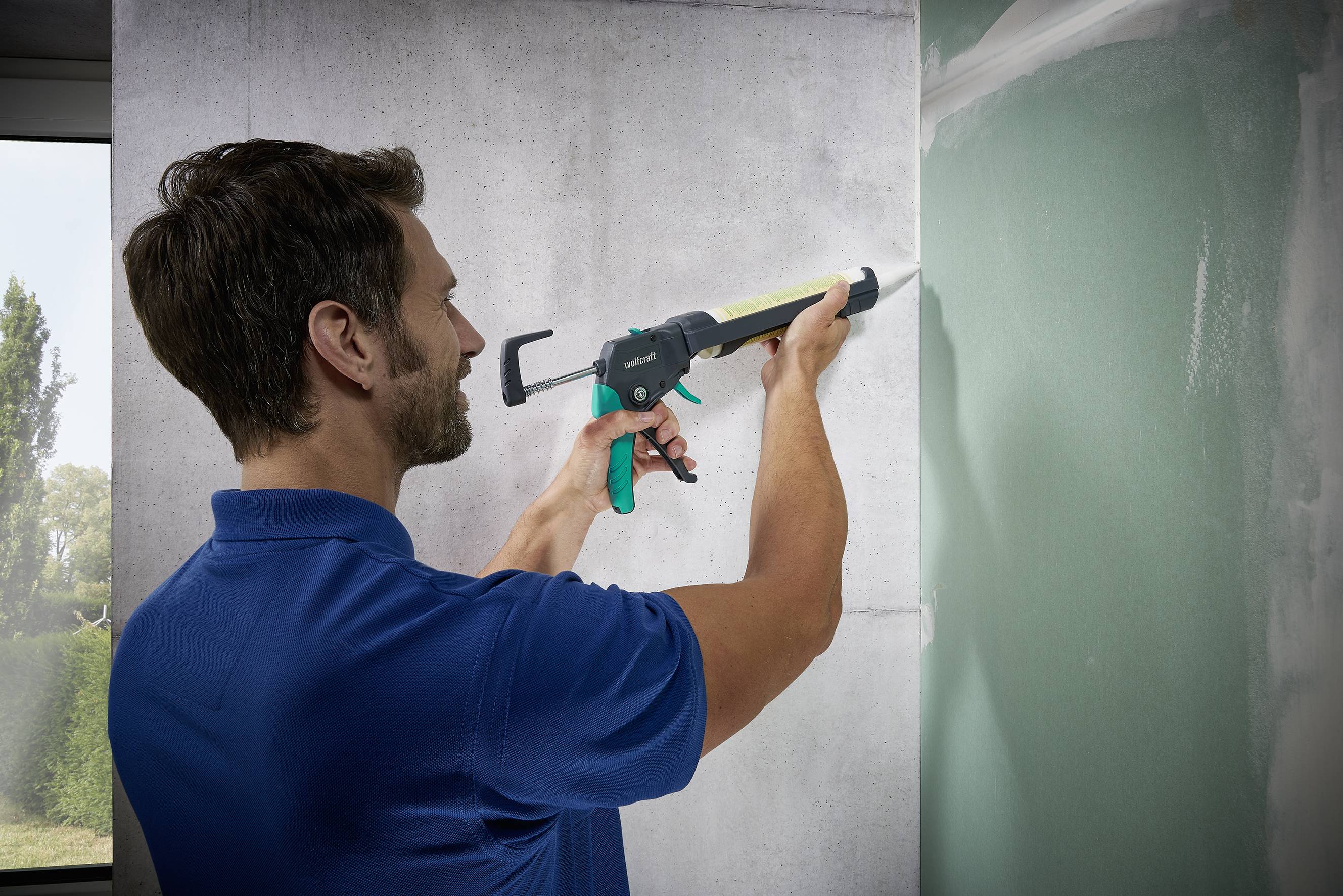 A man in a blue shirt is using a caulking gun to apply sealant to a wall. A window with trees is visible on the left.
