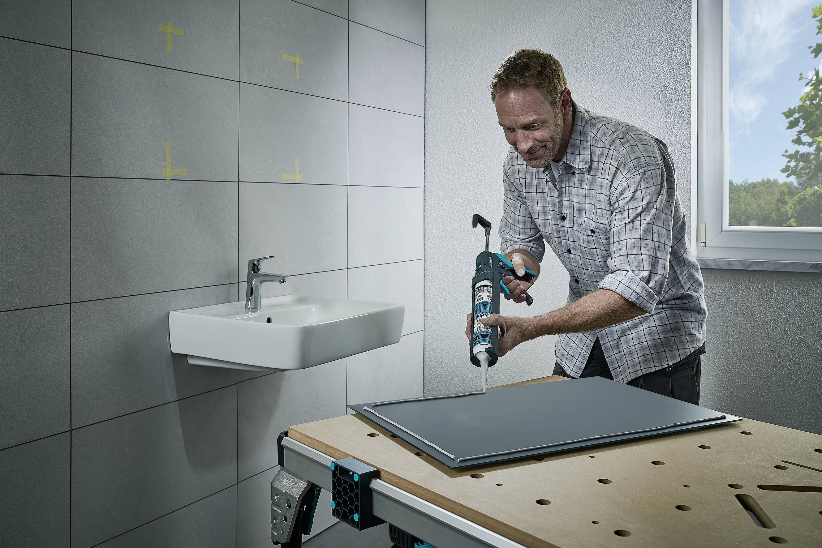 A man is applying glue to a plate placed on a table in a bathroom with a sink and tiled wall.