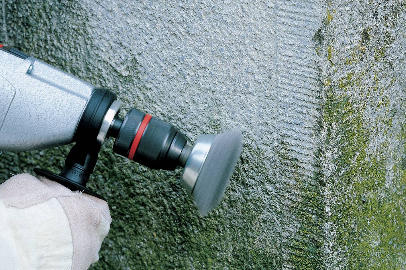 A hand holds a machine to grind a concrete wall. The wall shows signs of moss and ageing.