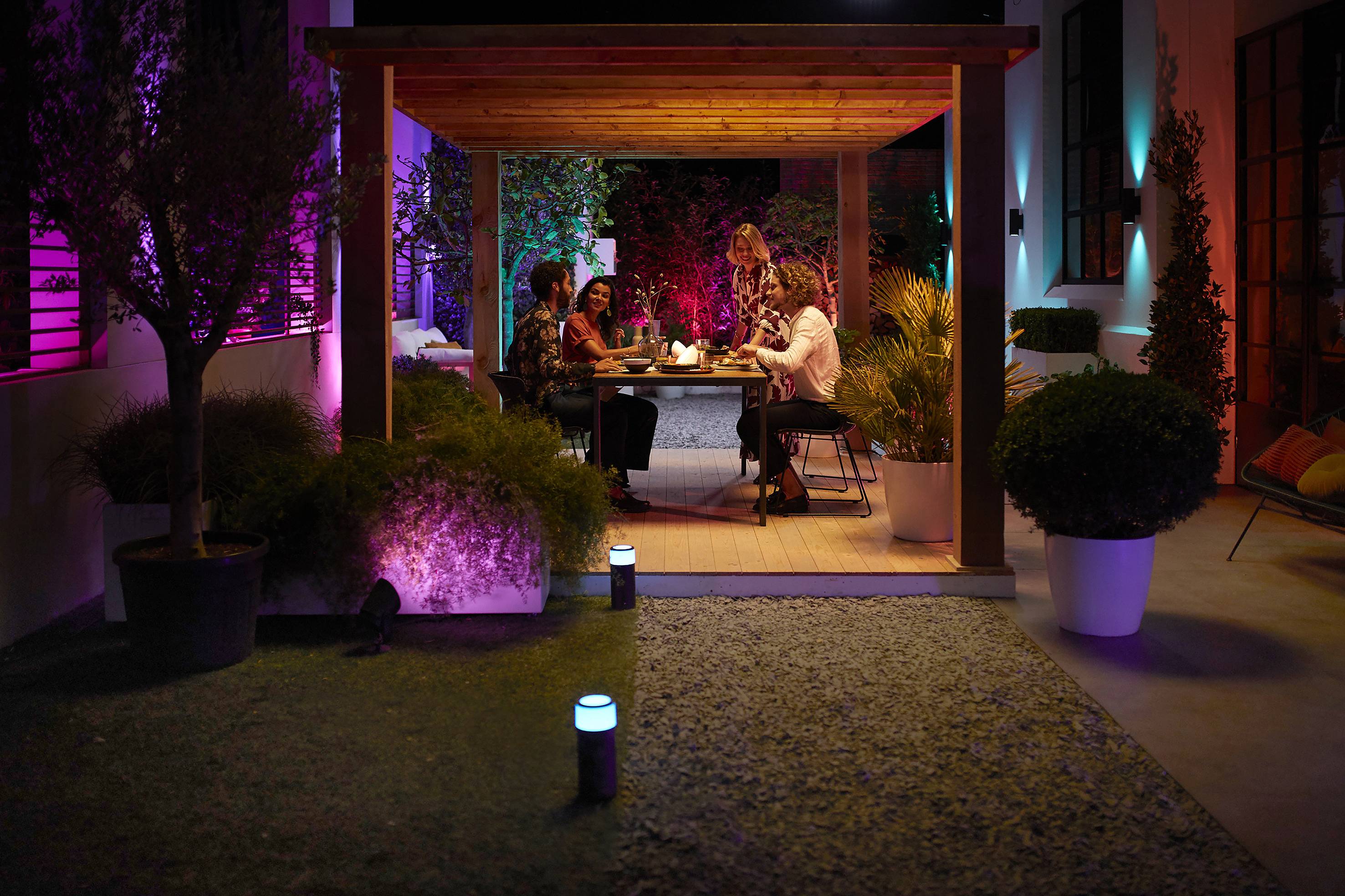 Four people are sitting at a table under a covered terrace, surrounded by colourful lighting and plants at night.