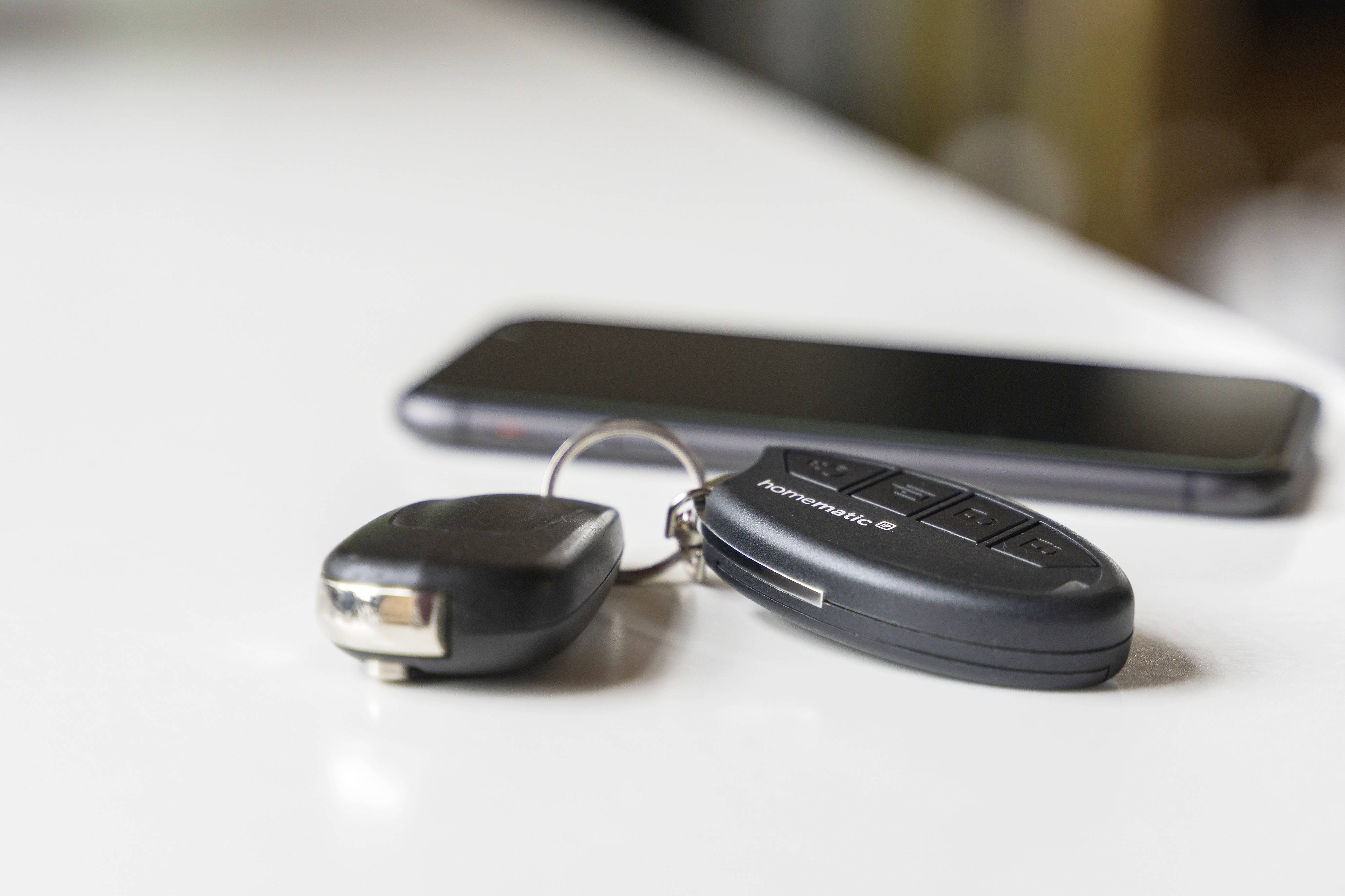 A car key and a smartphone are lying on a table. The focus is on the key, with the smartphone blurred in the background.