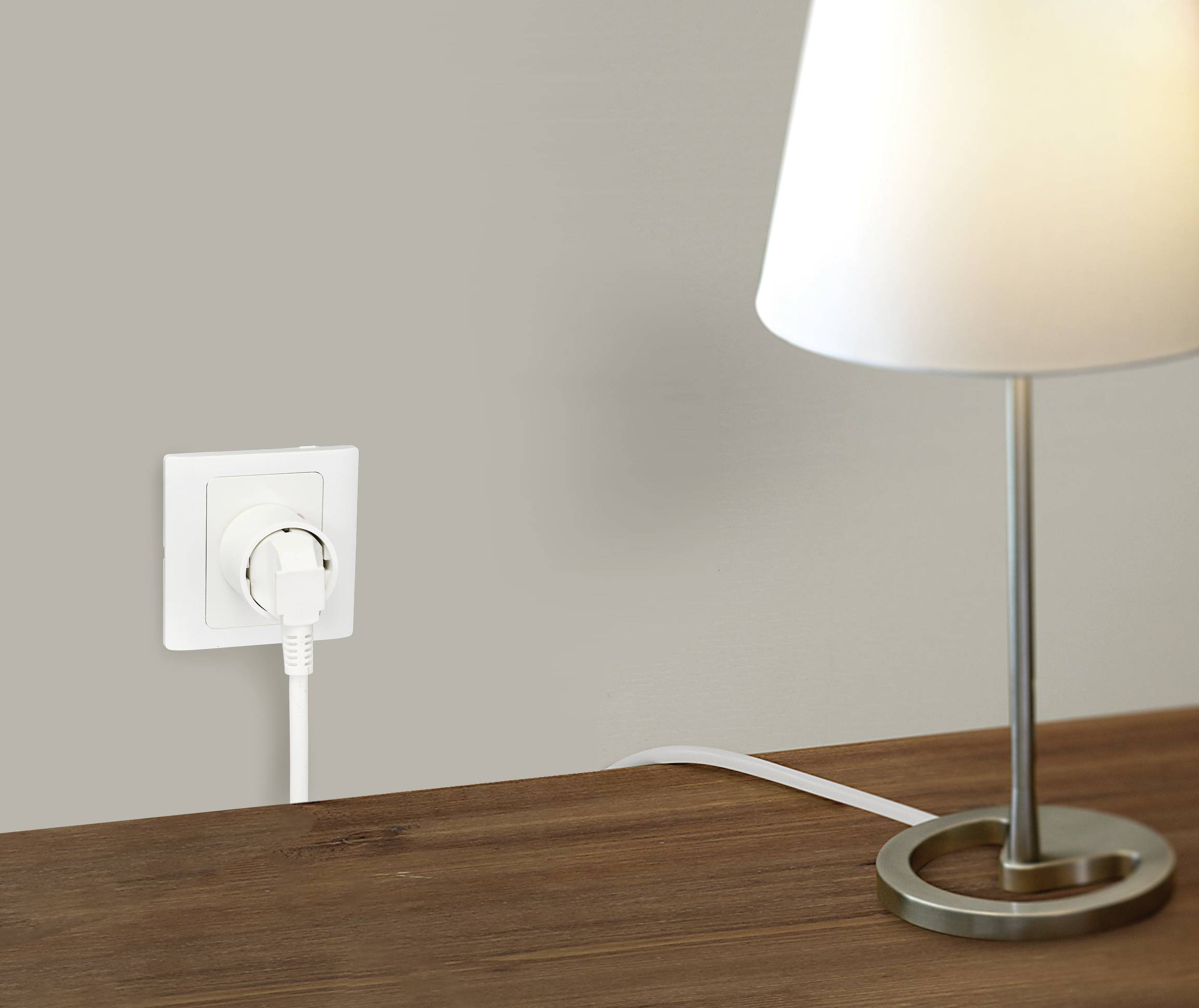 A table lamp illuminates on a wooden table. The lamp's cable is connected to a white wall socket.