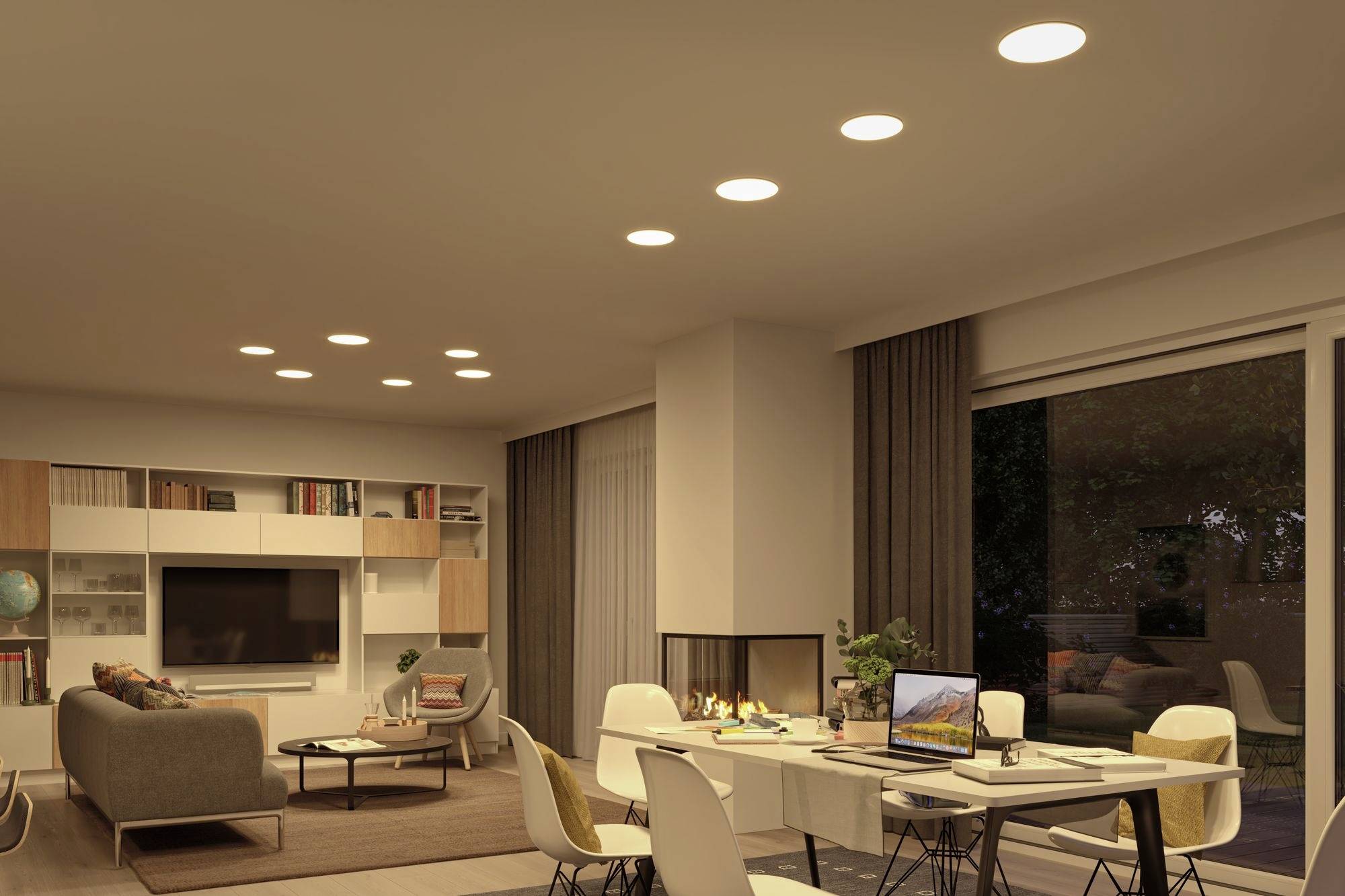 A modern living room with a dining area. The table is set with a laptop and books. Soft lighting creates a cosy atmosphere.