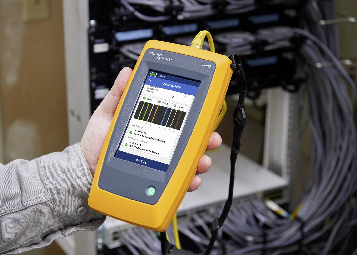 A person is holding a yellow network testing device in front of a server rack. Test results are displayed on the screen.
