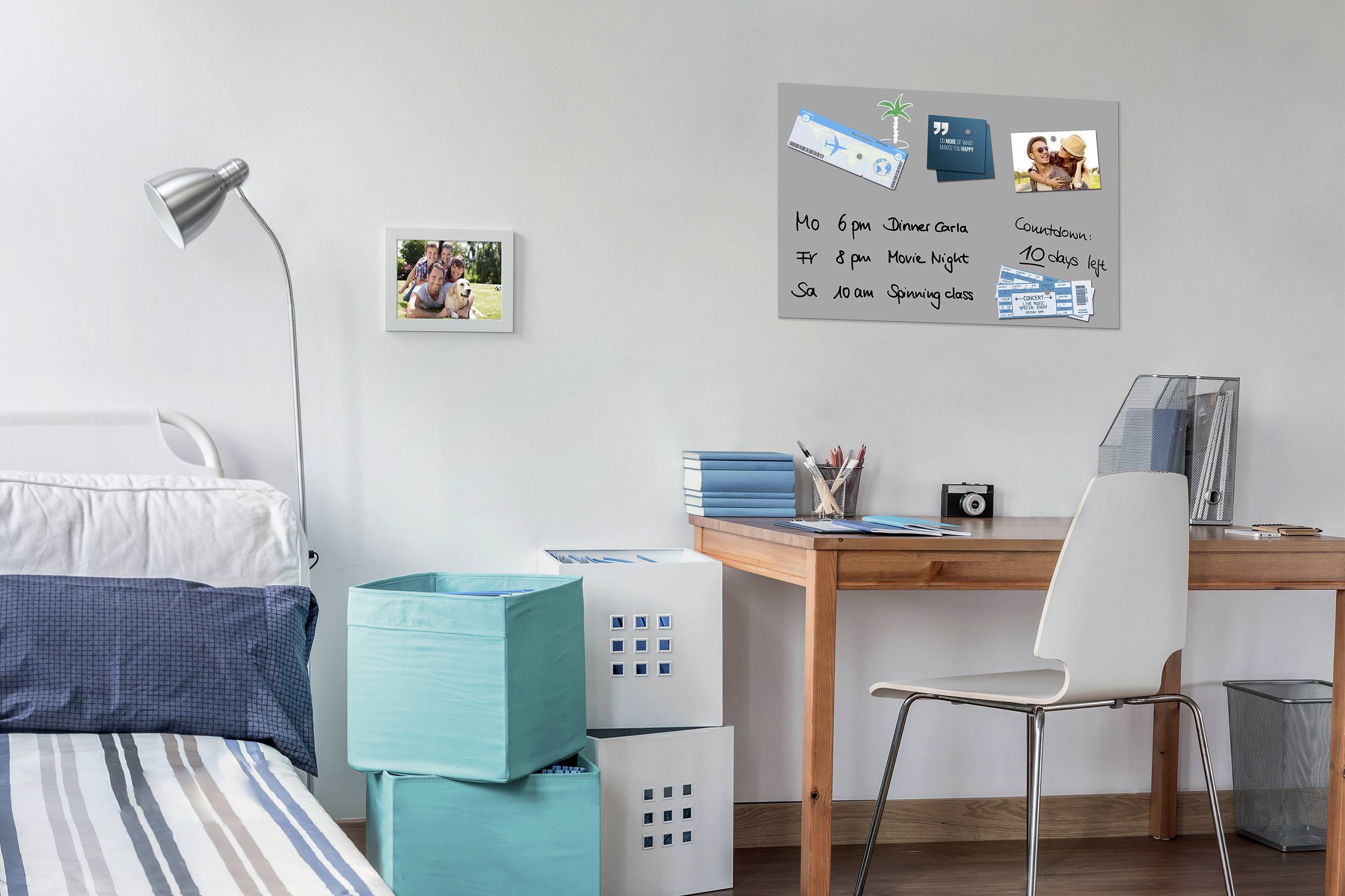A bedroom with a desk and a notice board featuring noted appointments. A bed with blue cushions and storage boxes beside it.