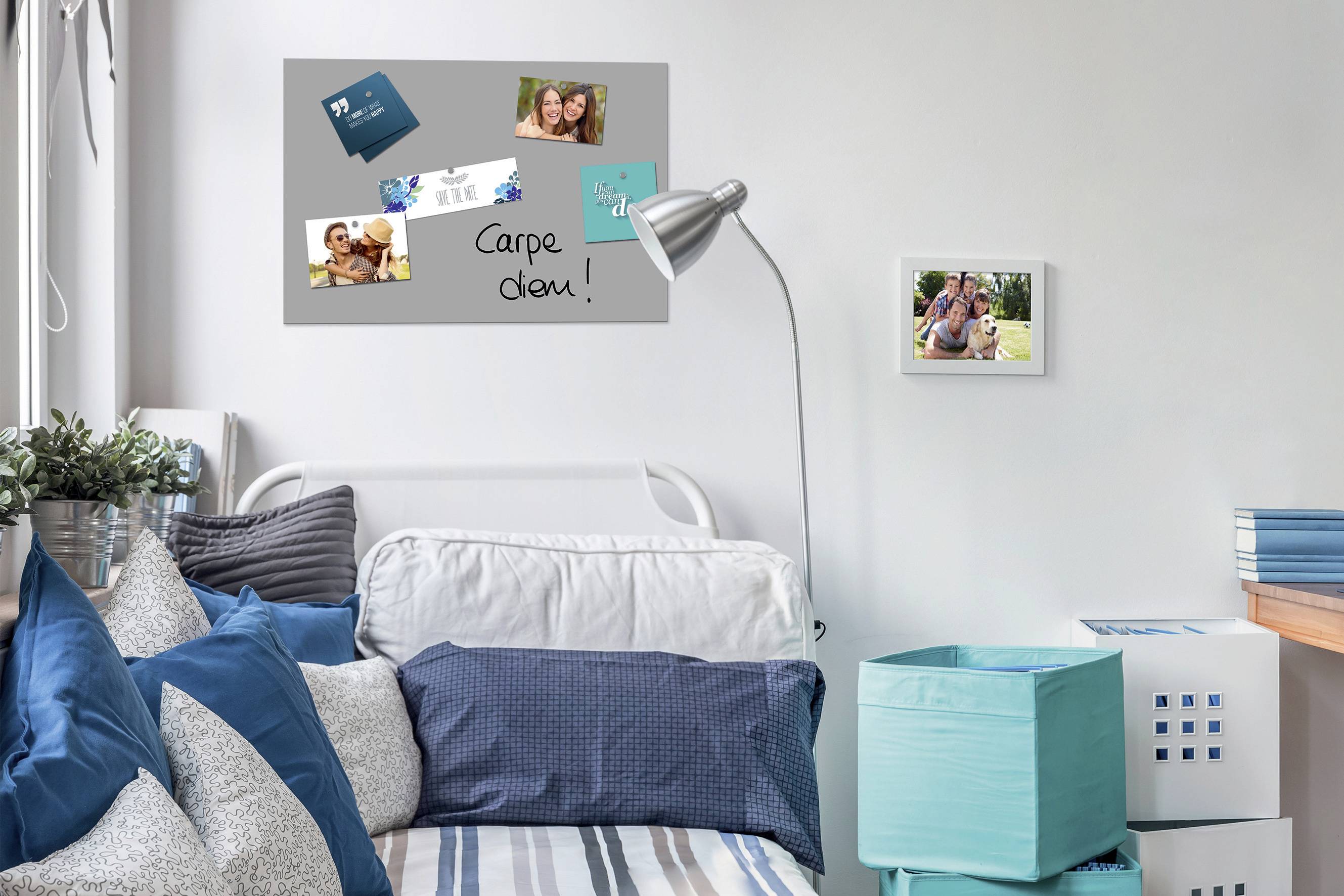 A bright bedroom with a bed full of blue cushions. On the wall hangs a pinboard with photographs and the inscription 'Carpe diem'.