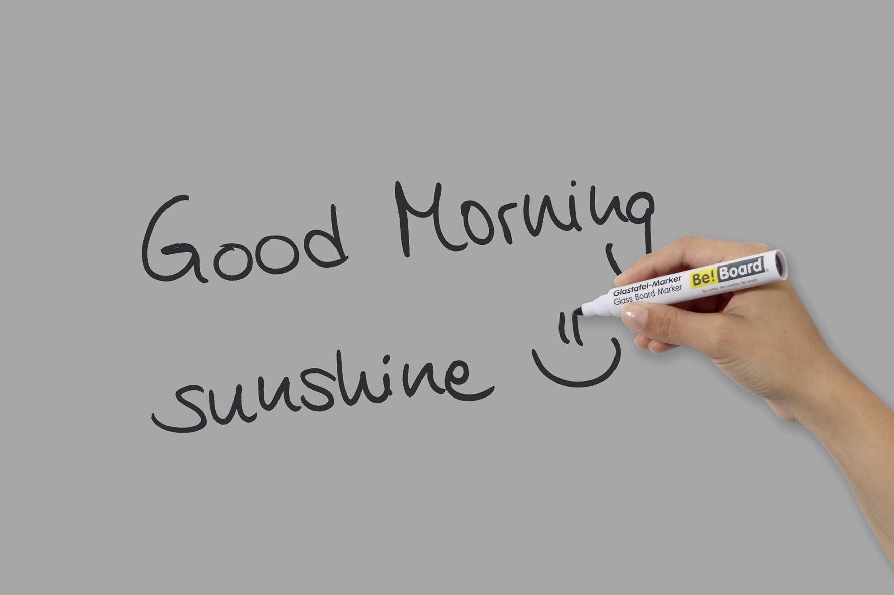 A hand writes with a pen on grey: 'Good Morning sunshine' and draws a smiley face.