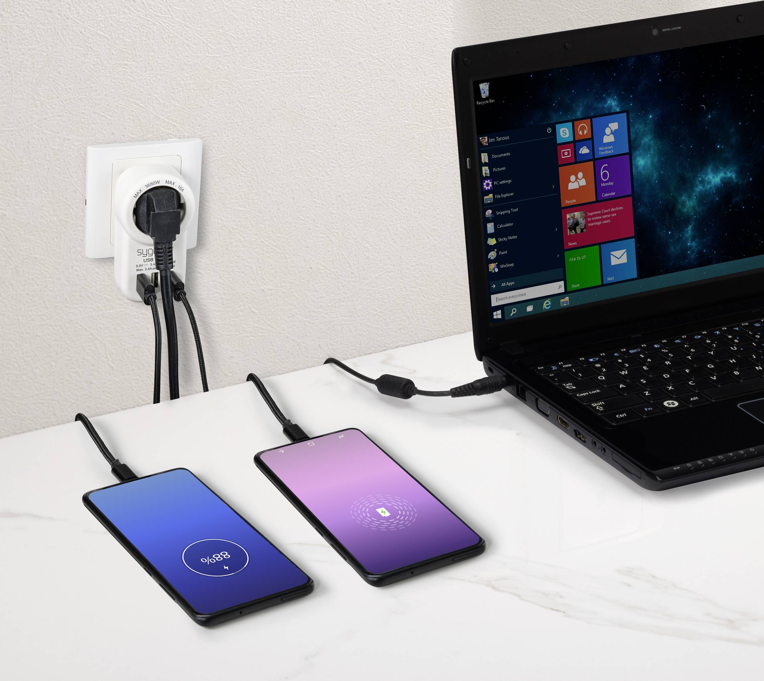 A black laptop with a Windows screen, two smartphones charging; one showing 90% battery, the other 18%, with a power socket in the background.