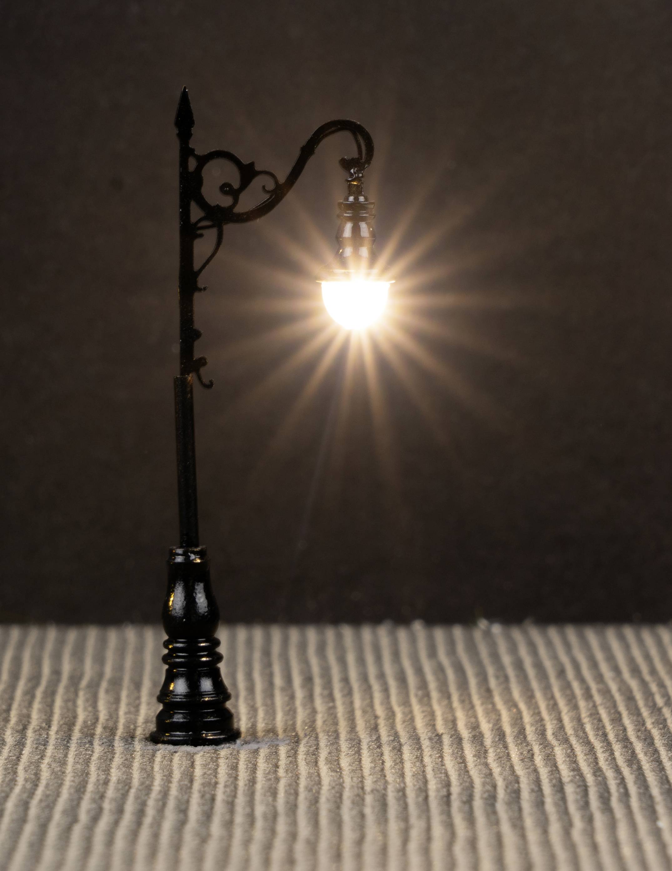 A small street lamp shines brightly, casting warm light onto the textured ground against a dark background.