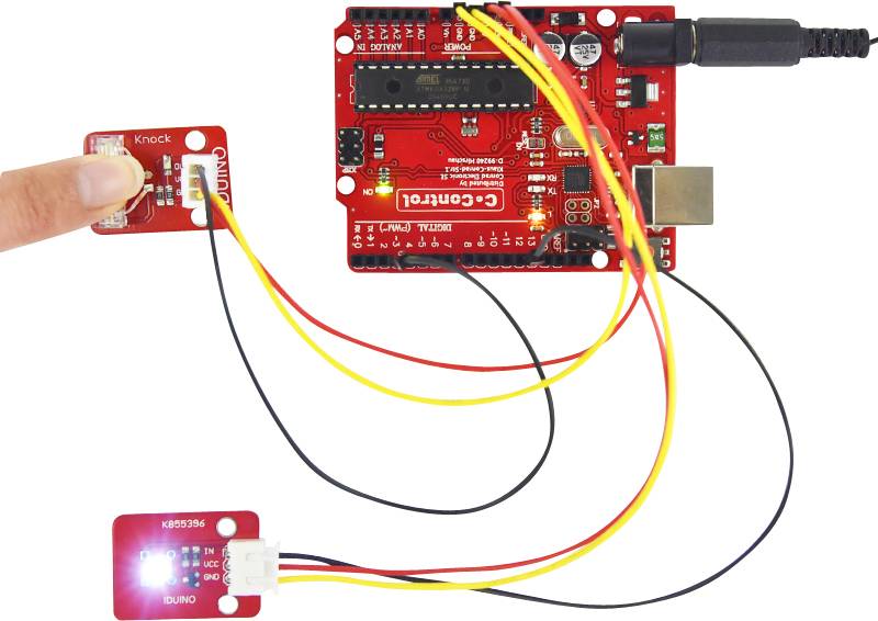A red microcontroller board is connected to a knock sensor and an illuminating LED module. A hand is pointing at the sensor.
