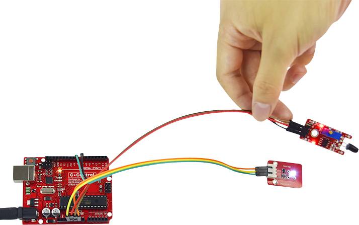 One hand holds a sensor that is connected via cables to a red microcontroller. Another sensor is also connected.