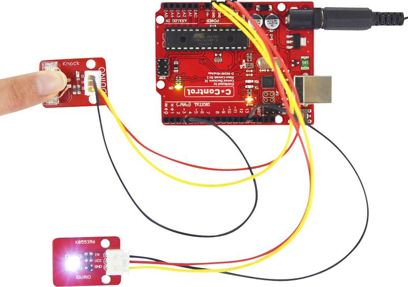 Red open-source circuit board connected with cables. A blue LED light and a sensor are connected to the circuit board.