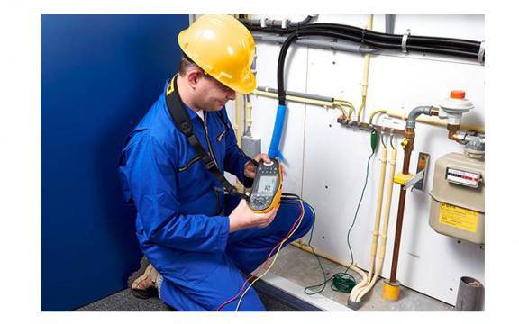 A technician in blue workwear and a yellow hard hat is using a measuring device to inspect pipes and conduits in a technical room.
