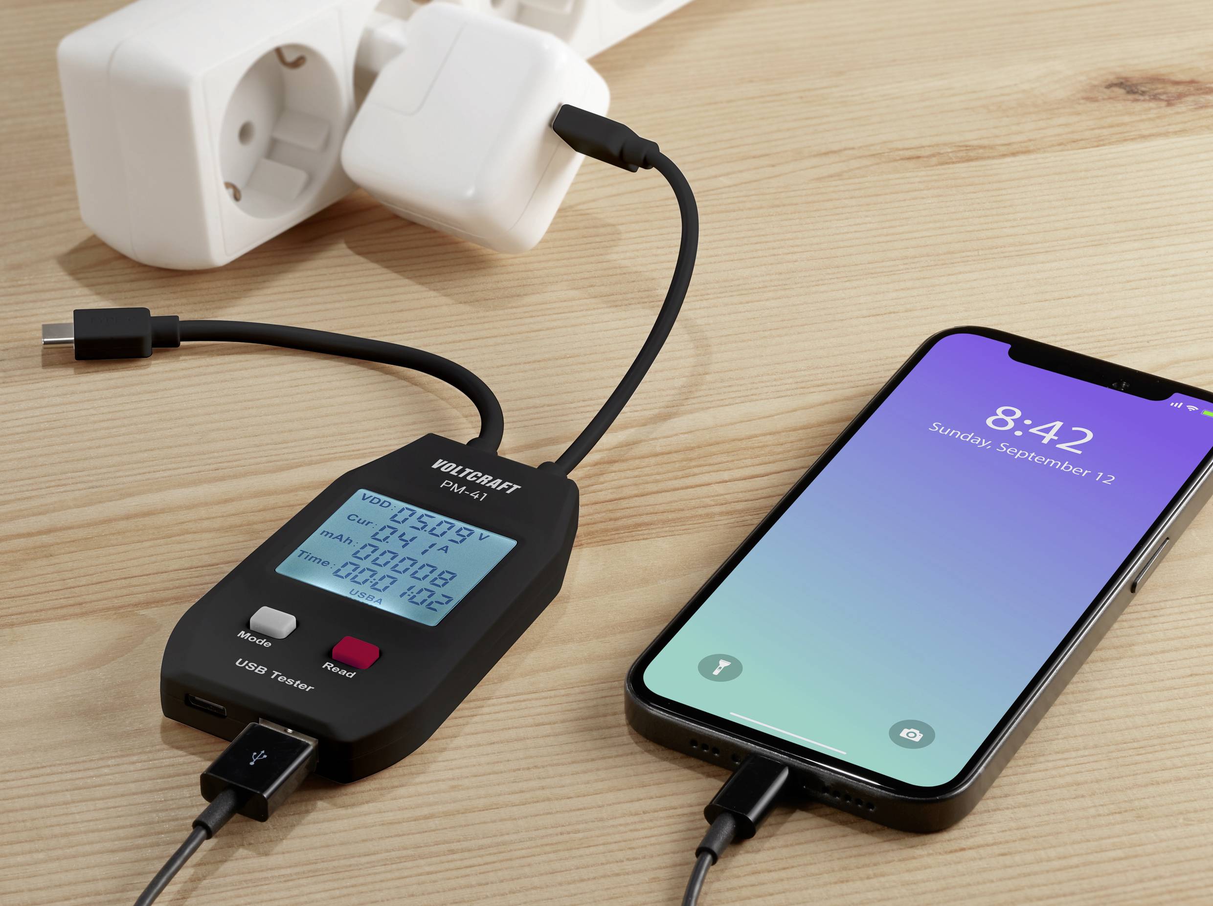A smartphone is being charged via a USB charging cable, connected to a measuring device that displays voltage, current, and time.