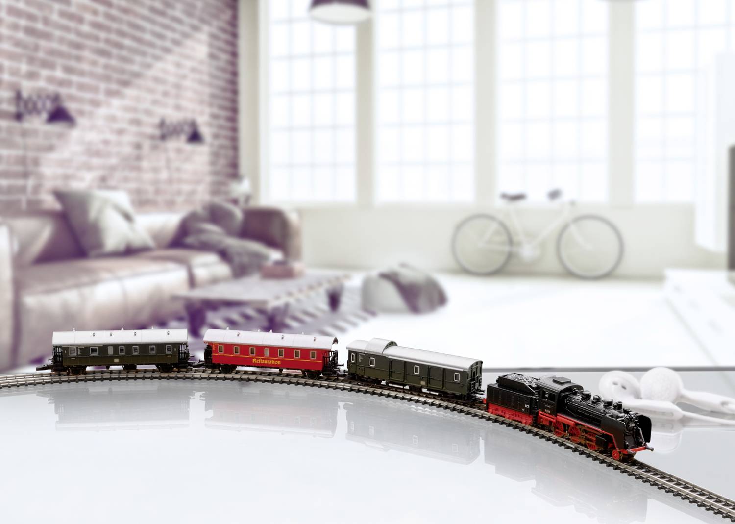 A model railway travels along a track system in front of a cosy living room with a brick wall and a bicycle in the background.