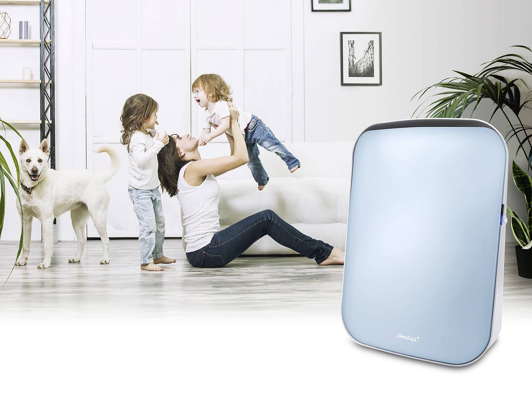 A woman is sitting on the floor, lifting a child up, while another child stands beside her. A dog is sitting to the left. An air purifier stands to the right.