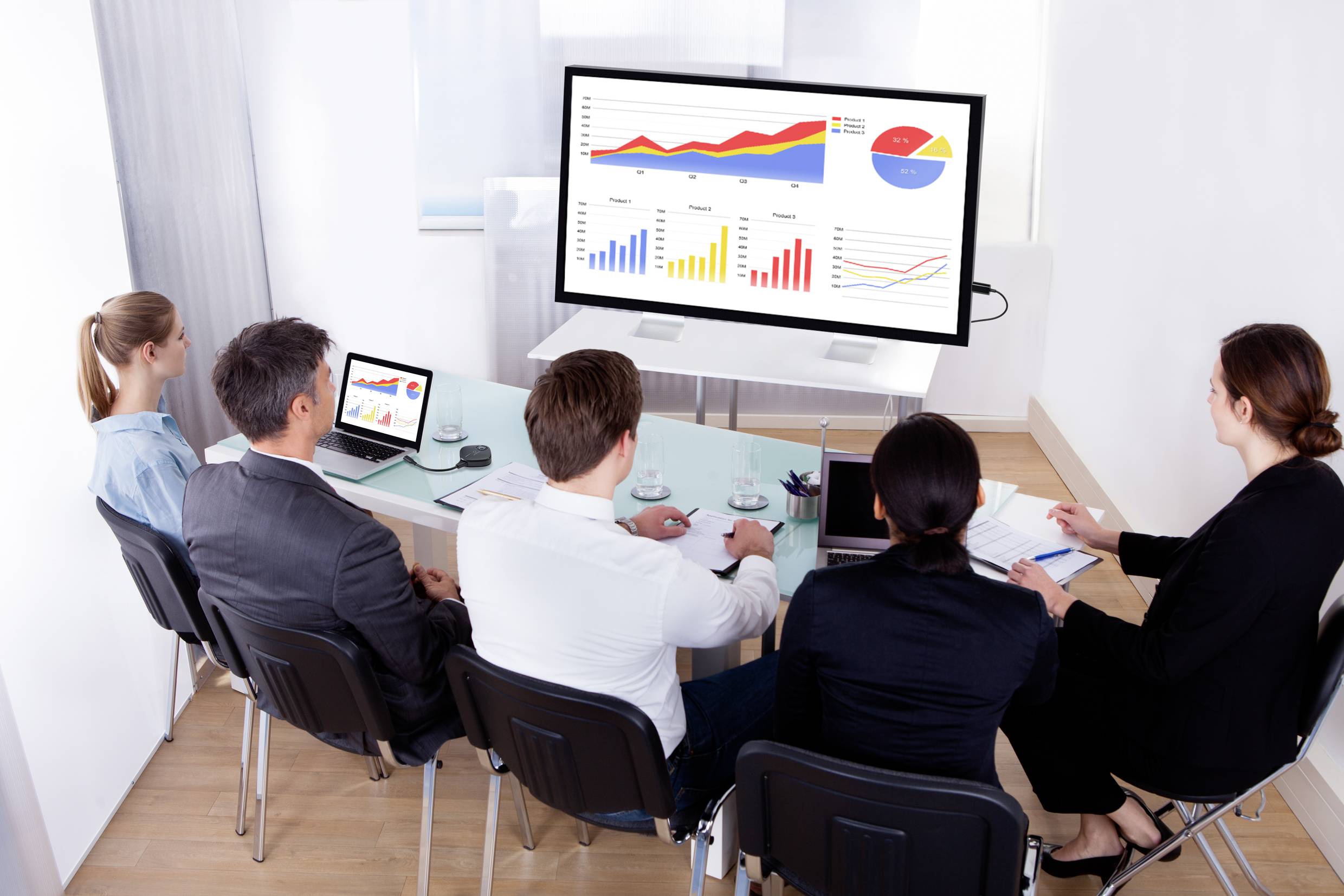 A group of people are sitting around a conference table, examining diagrams and graphs on a large screen.