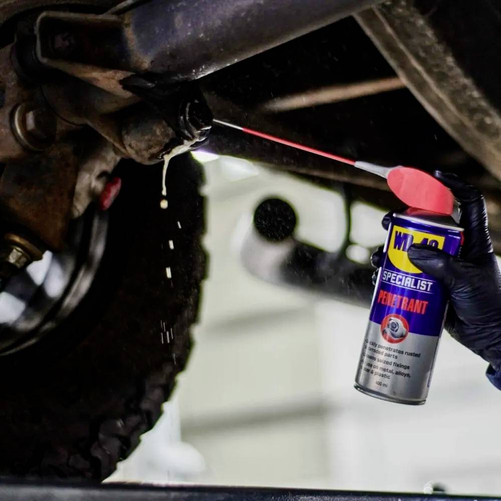 A person is applying a lubricant to a car part to remove rust. They are holding a spray can with the label 'WD-40 Specialist'.