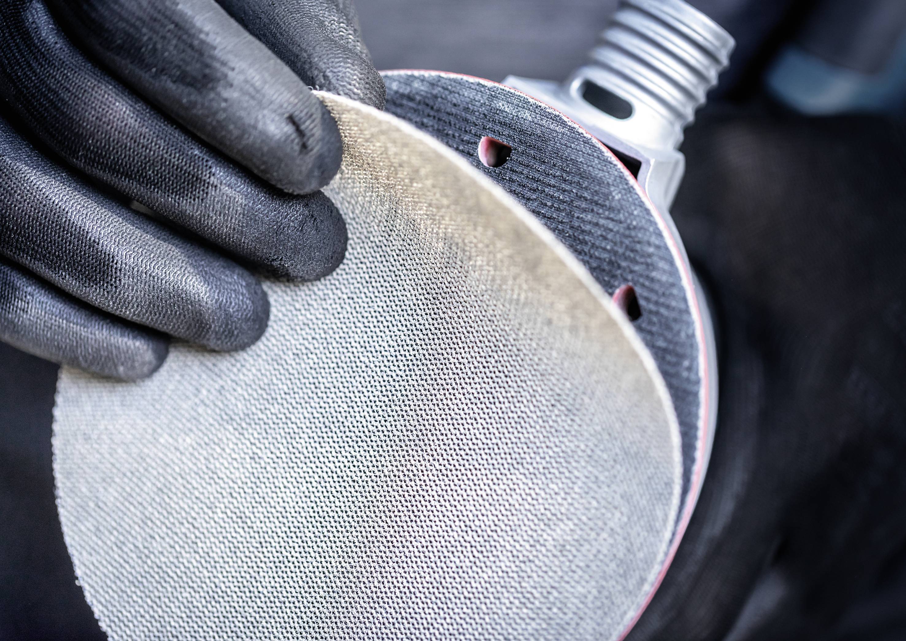 A person is wearing black gloves and holding a round piece of sandpaper. It is being placed on a sanding disc, ready for sanding.