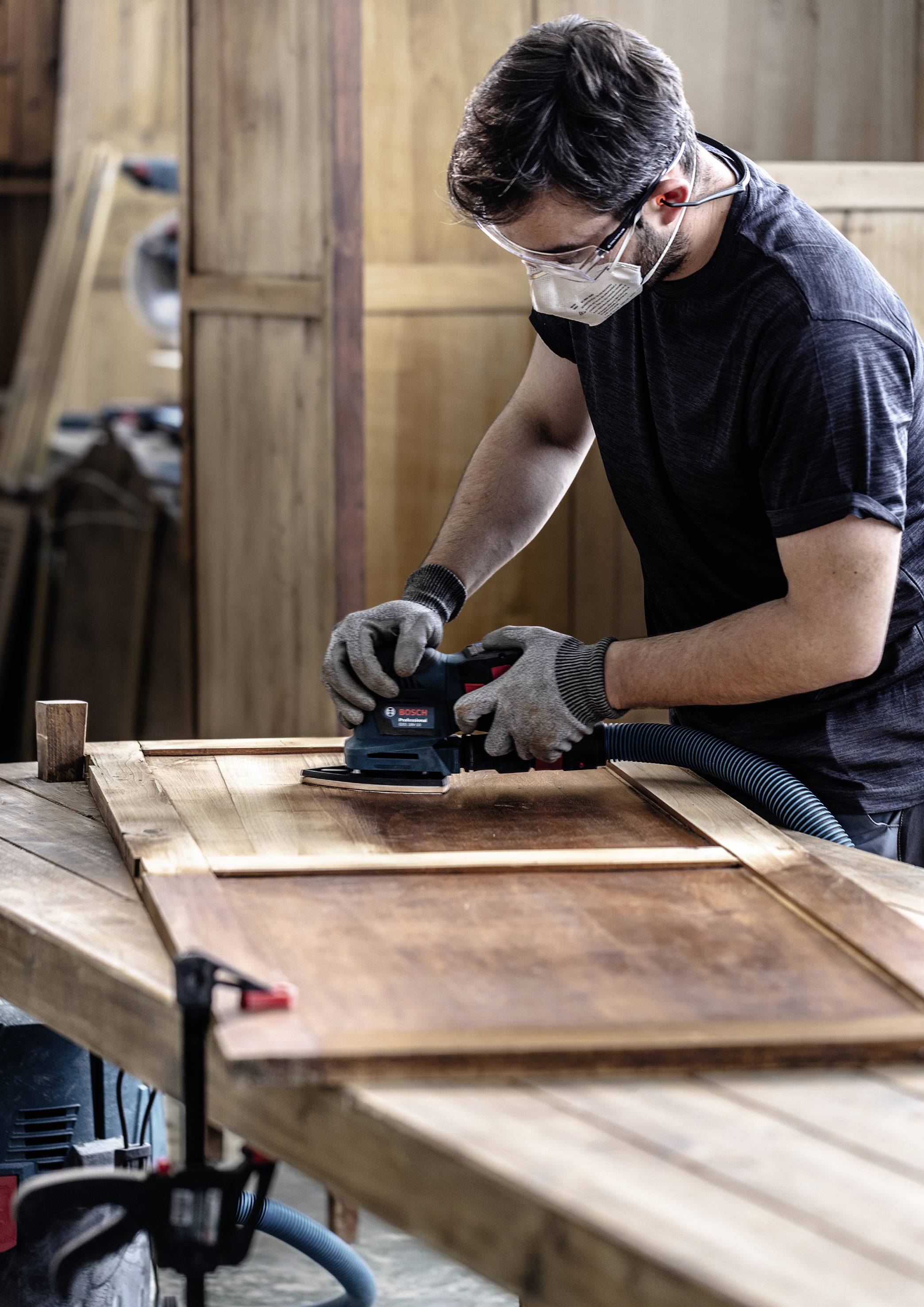 A person is sanding a wooden door in a workshop. They are wearing a protective mask and gloves. Wood shavings are visible on the table.