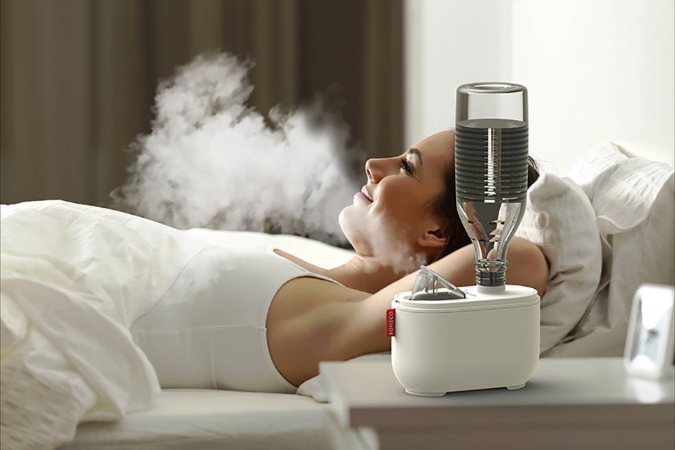 A woman is lying relaxed in bed, inhaling the vapour from a humidifier beside her.