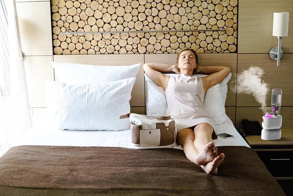 A woman in a white dress is relaxing on a hotel bed. Next to her is a bag and a running humidifier.