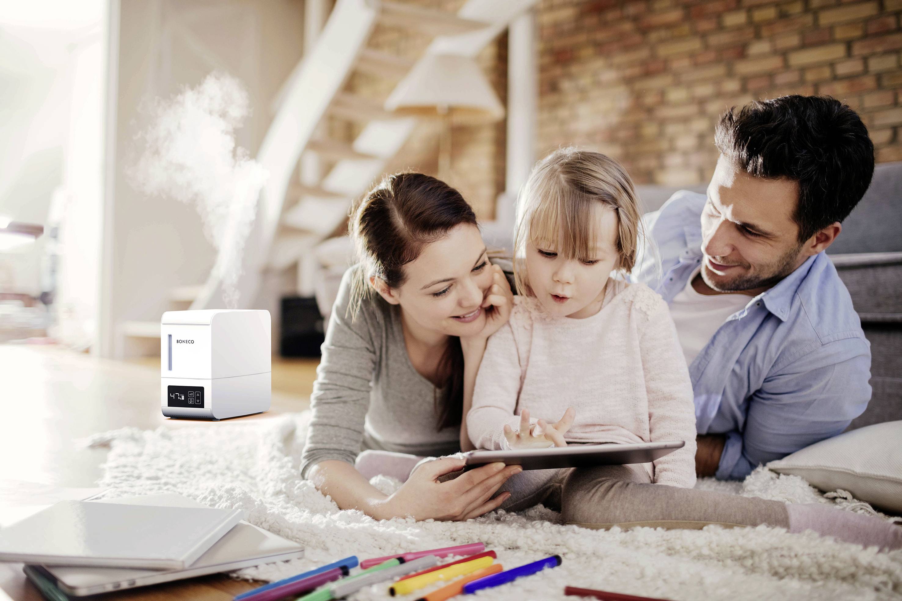 A child is lying with their parents on a rug, drawing on a tablet. A humidifier stands in the background.