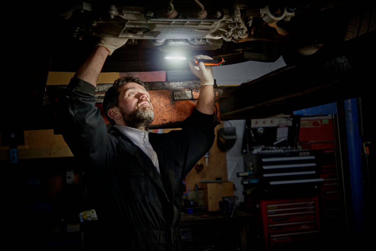 A mechanic is working in a workshop underneath a raised vehicle and illuminating the area with a lamp to inspect parts.