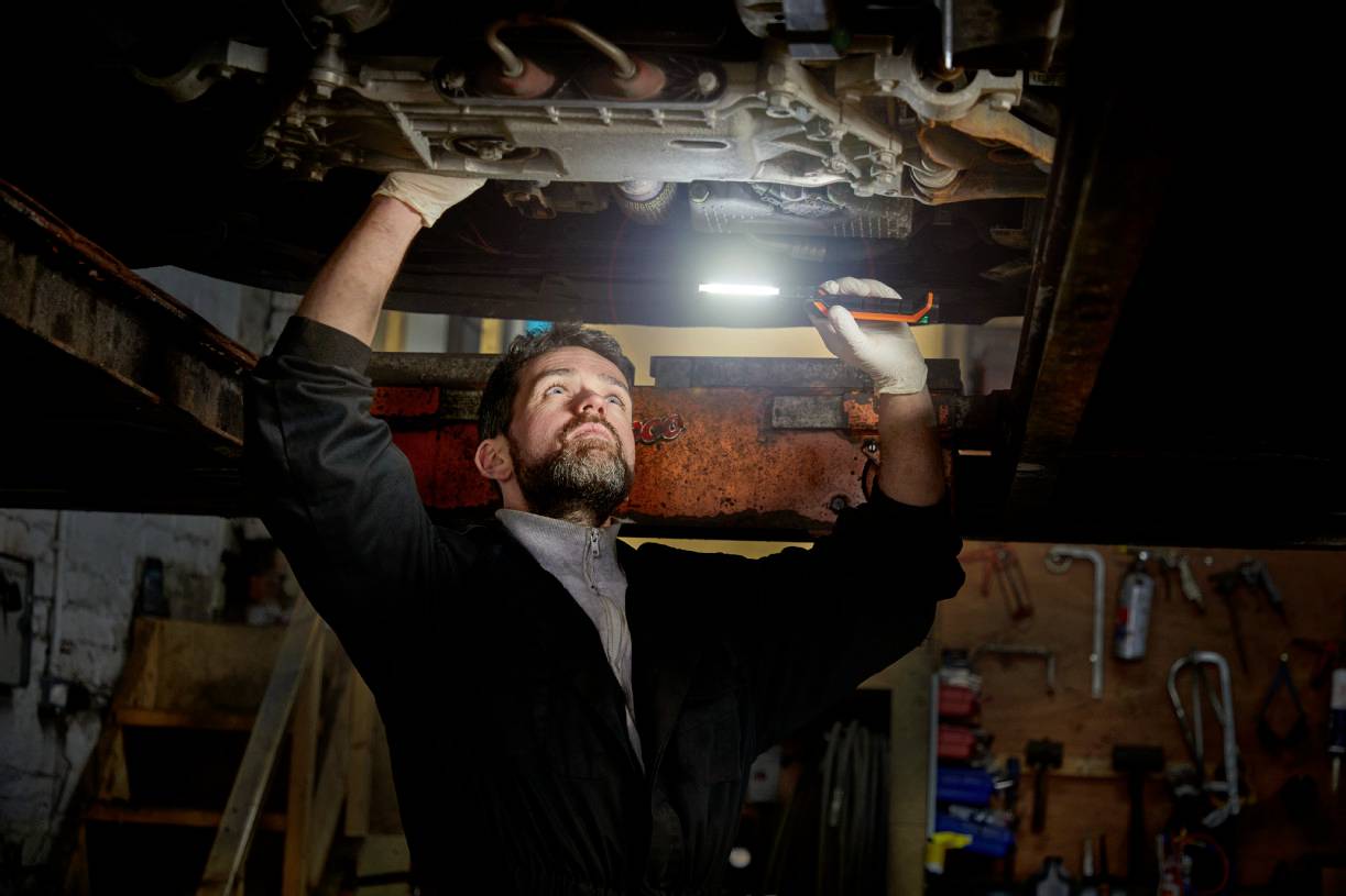 A mechanic beneath a car in a workshop, focused and holding a torch in his hand while carrying out repairs.