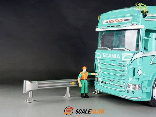 A model lorry is parked beside a crash barrier section. A figure in safety clothing is touching the front bumper of the lorry.
