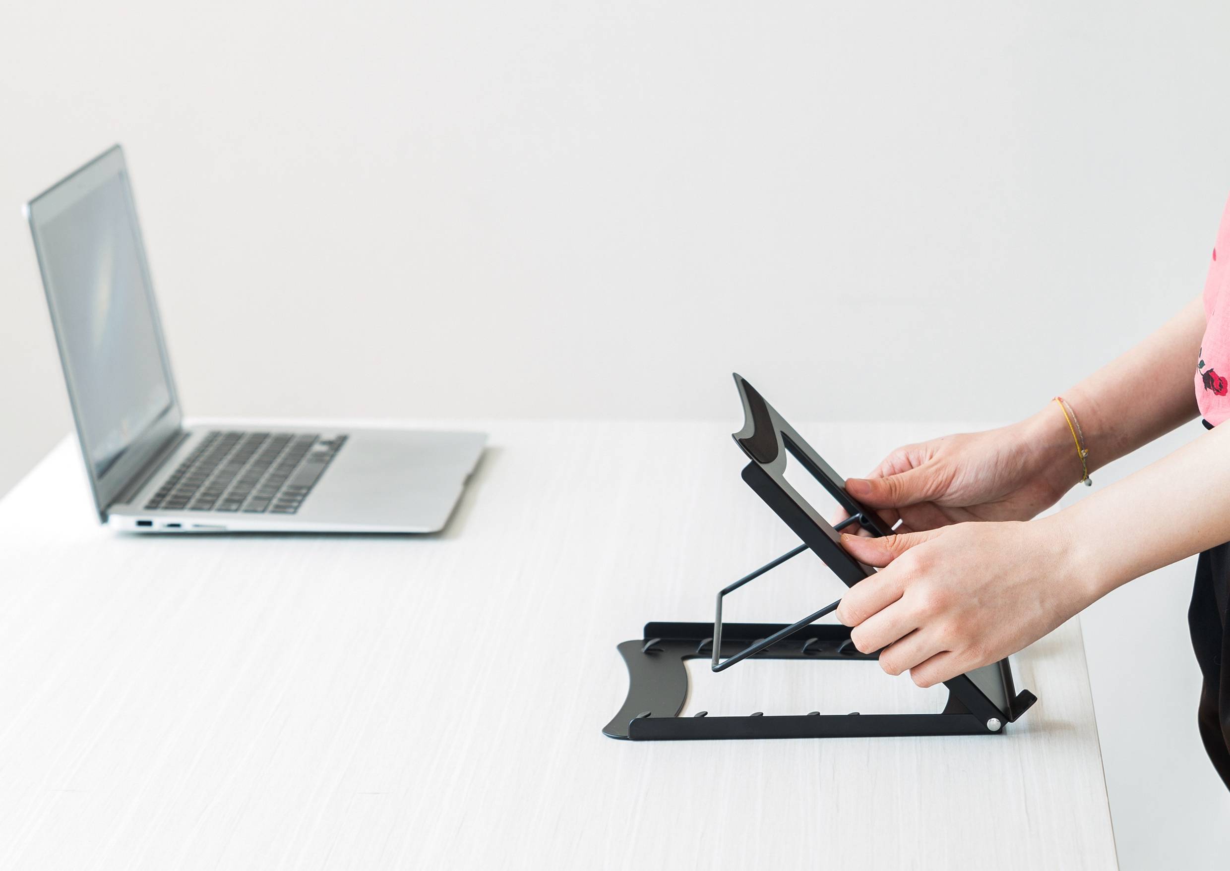 A person is positioning a black laptop stand on a white table, next to an open laptop.