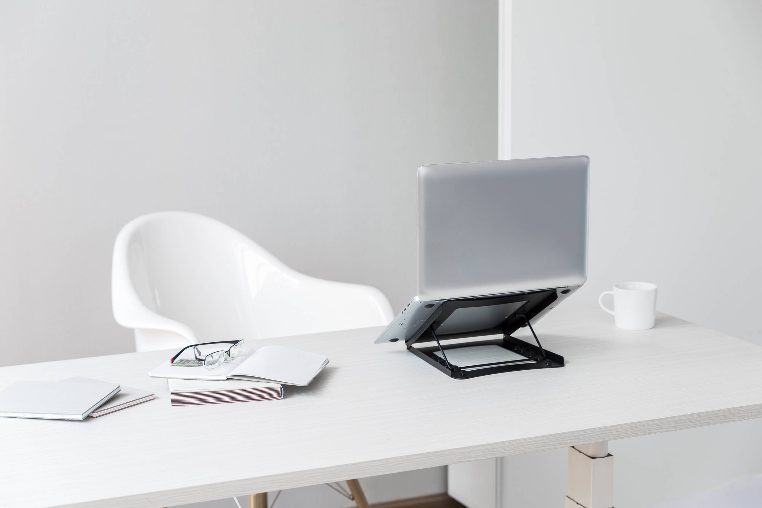 A modern, empty desk with an open laptop on a stand, a notebook, and a pair of glasses. A white chair stands in the background.
