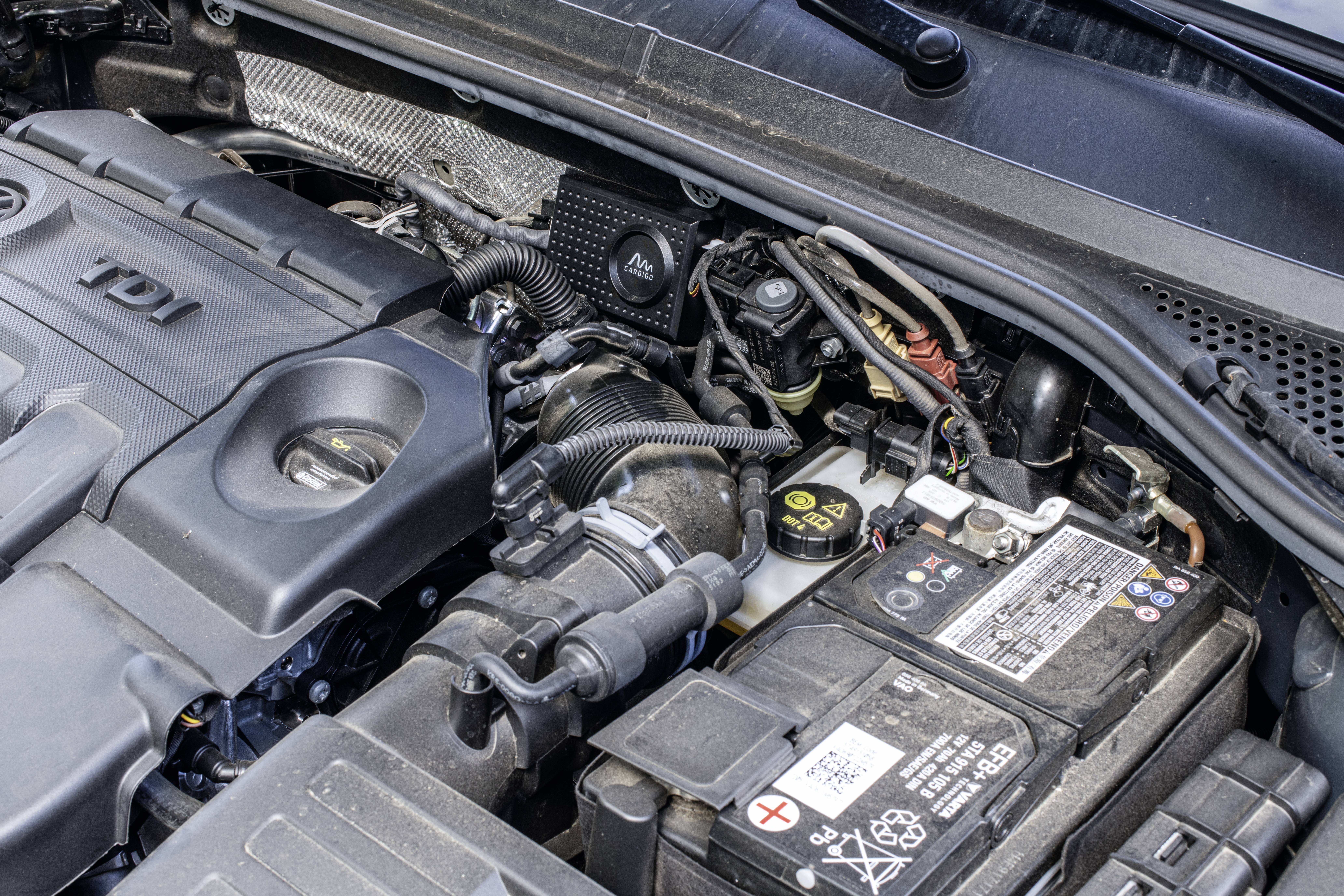 Engine bay of a vehicle, showing components such as car battery and engine cover. Wiring and hoses are clearly visible.