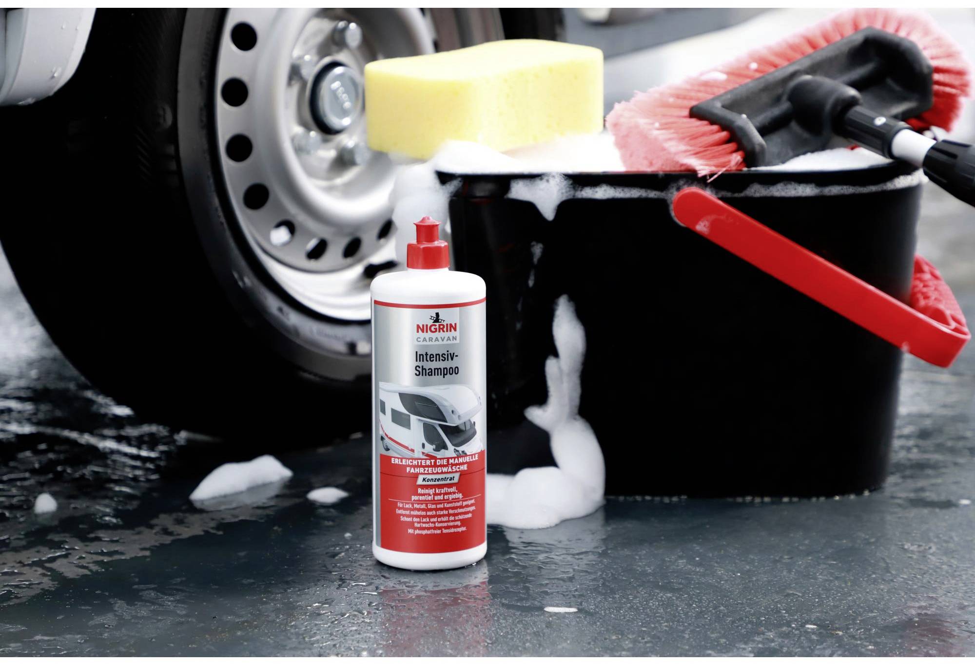 A bucket with a red brush and yellow sponge is positioned next to tyres. A bottle of 'Intensive Shampoo' by Nigrin is in the foreground. Foam is visible.