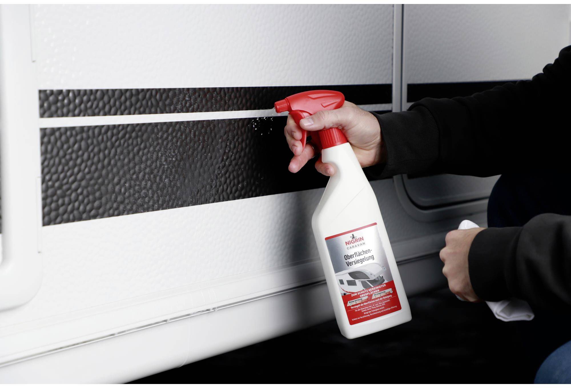A person is spraying a cleaning solution from a white bottle with a red label and spray nozzle onto the surface of a vehicle.