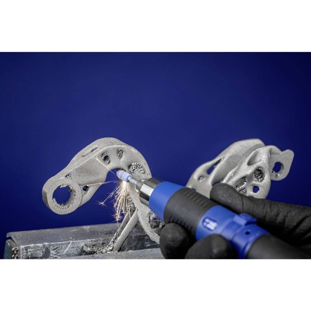 A 3D-printed metal component is being machined with a blue hand tool, with sparks flying. Hands wearing black gloves are holding the tool.