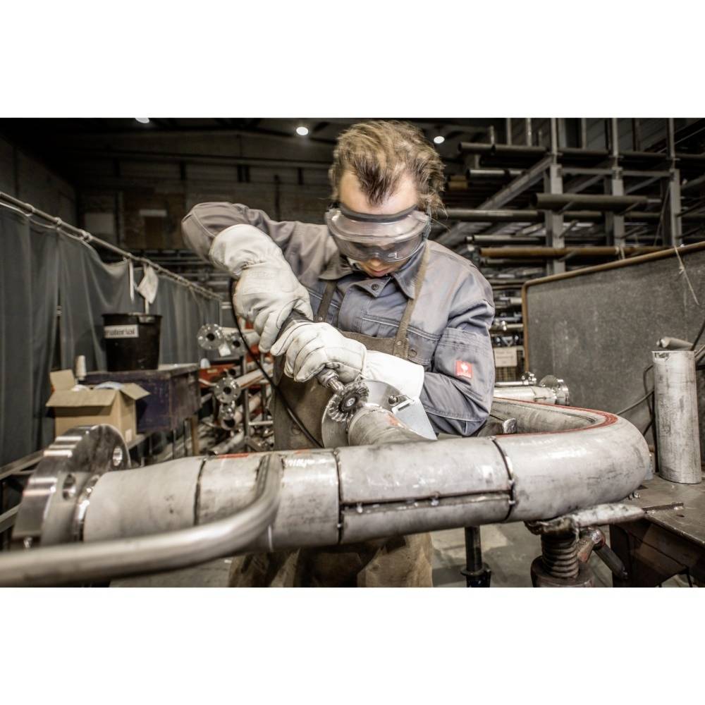 A worker wearing safety glasses and gloves is processing metal pipes in a workshop environment, surrounded by tools and equipment.
