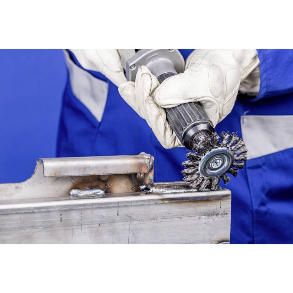 A person in blue workwear is using a rotating wire brush to polish metal. The scene depicts precision work.