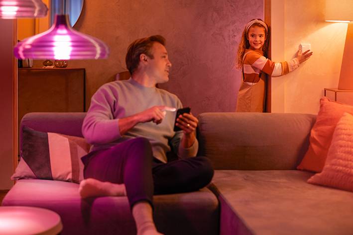 A man is sitting on a sofa with a smartphone, whilst a girl presses a light switch on the wall. The room is atmospherically lit.