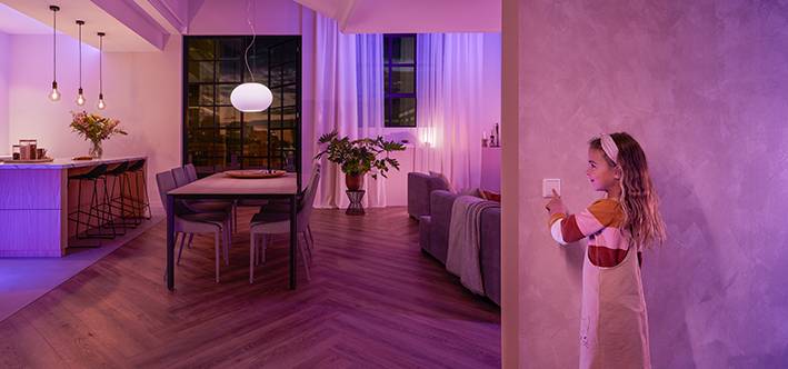A child stands by a light switch, looking at a cosily lit living room with a dining table, chairs, and plants.