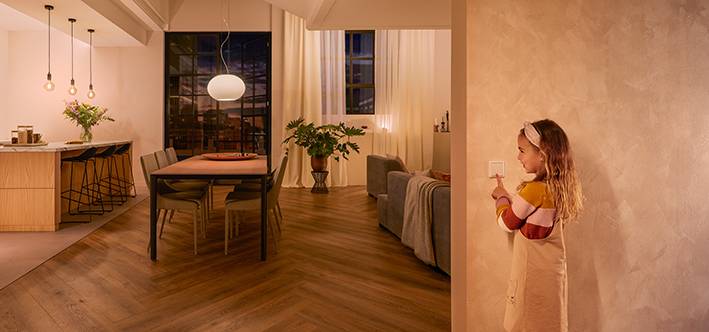 A child operates a light switch and illuminates a modern, cosy living and dining area with warm light in the background.