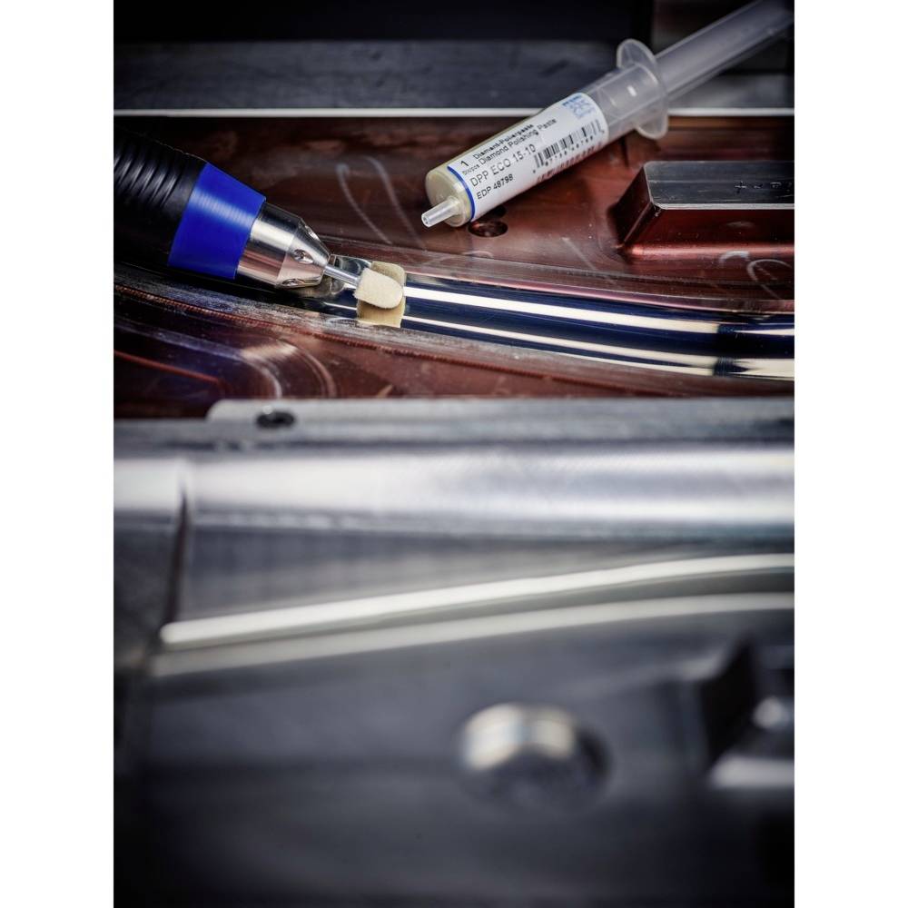 A soldering iron touches a small, beige component on a metallic surface. In the background, a solder paste syringe lies ready.