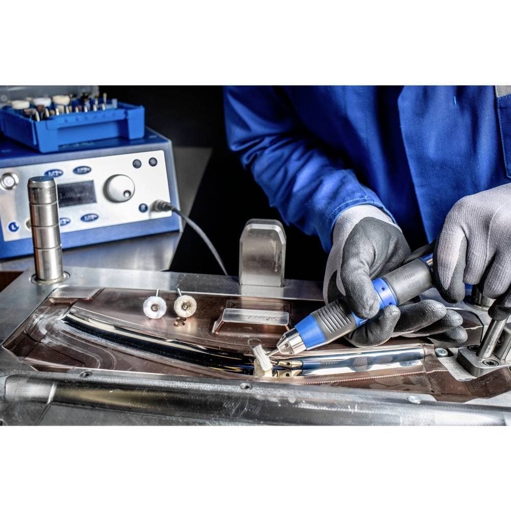 A person wearing blue protective clothing and grey gloves working on a metalworking machine with various tools.